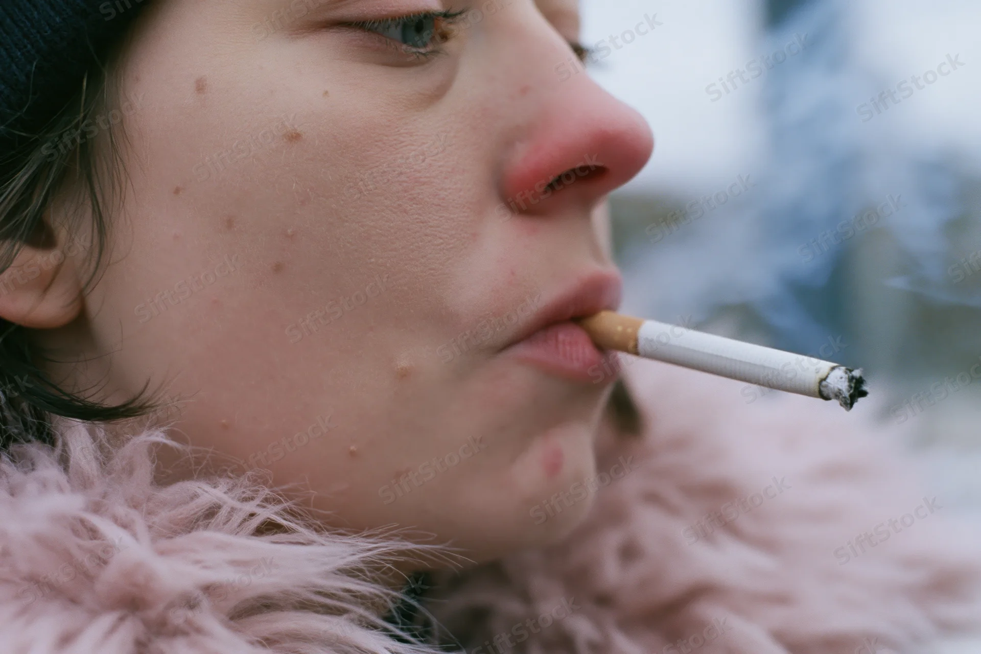 Young woman in profile smoking a cigarette outdoors in winter, wearing a pink fur coat and black beanie