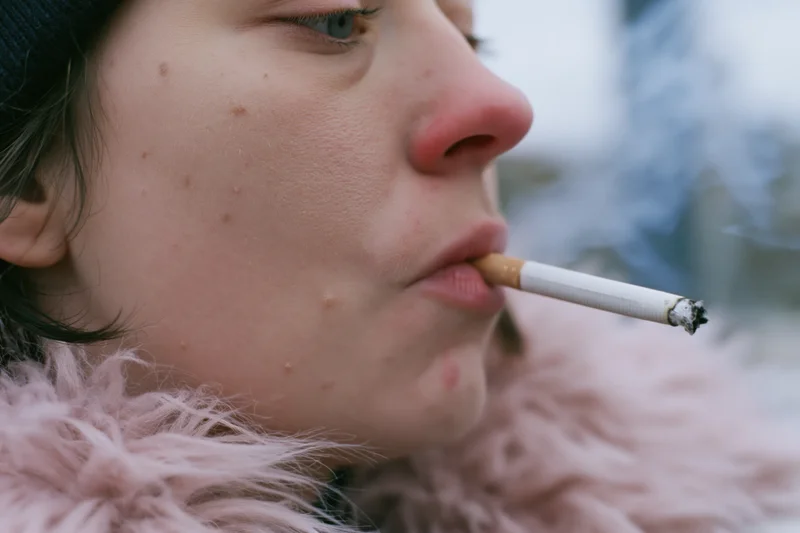 Young woman in profile smoking a cigarette outdoors in winter, wearing a pink fur coat and black beanie