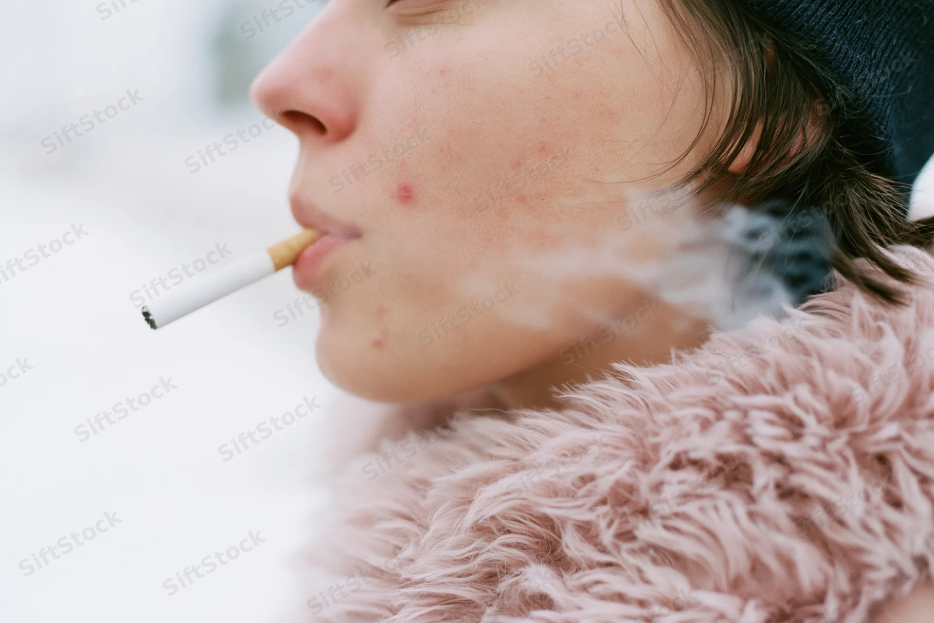 Young woman in profile smoking a cigarette outdoors in winter, exhaling smoke, wearing a pink fur coat and dark beanie