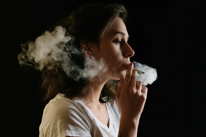 Young woman in profile exhaling smoke from a cigarette against a dark background