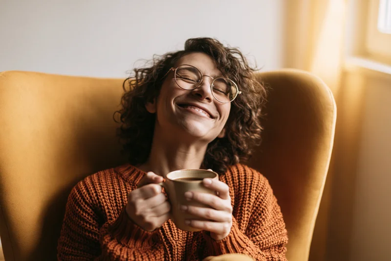 Woman laughing with eyes closed and head tilted back while holding a mug in a cozy armchair by a sunlit window