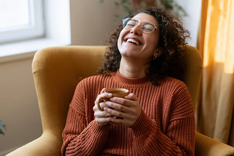 Woman laughing with eyes closed and head tilted back while holding a mug in an armchair indoors