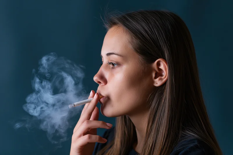 Young woman in profile smoking a cigarette with exhaled smoke against a dark blue background