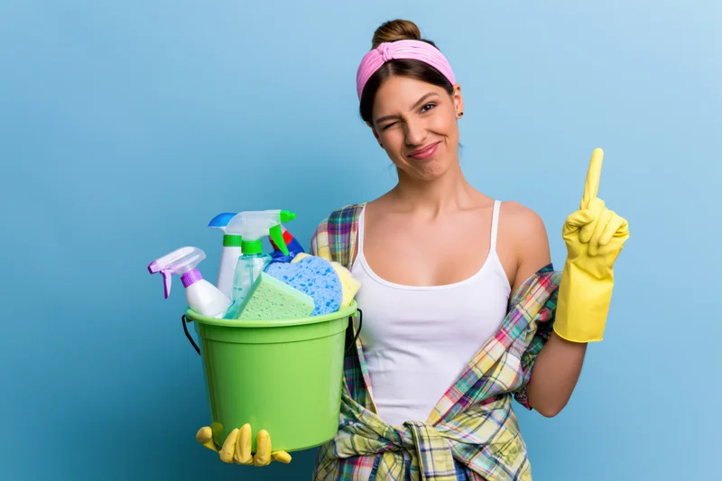 Woman winking and pointing while holding a green bucket of cleaning supplies against a blue background