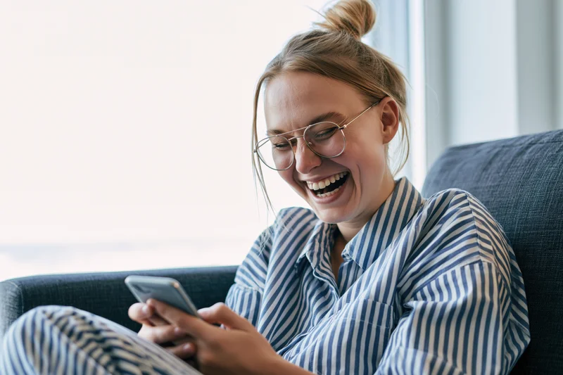 Young woman laughing while using smartphone on couch at home