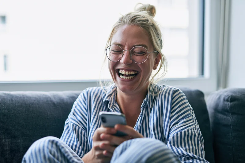 Woman laughing while using smartphone on couch at home