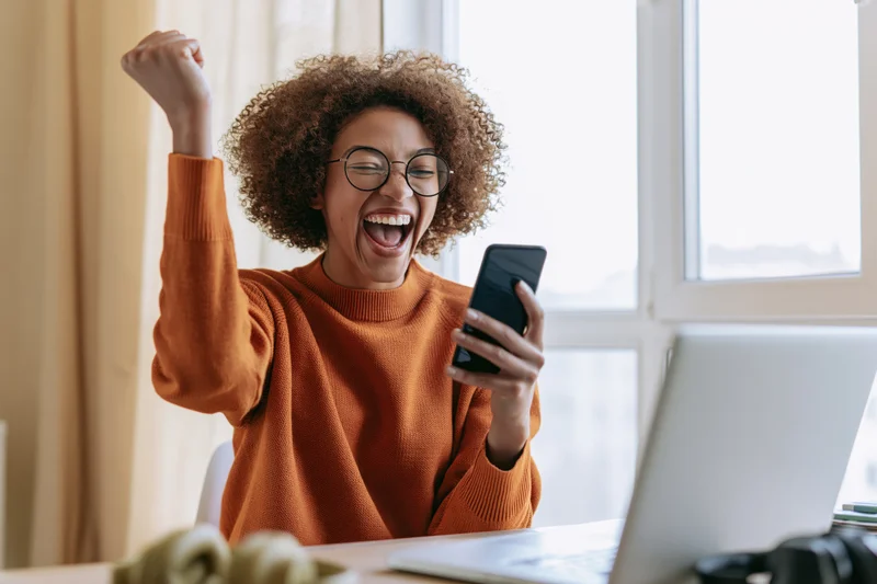 Woman celebrating with raised fist while holding smartphone at home office desk
