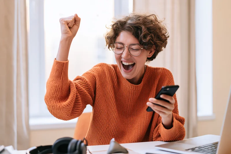 Woman celebrating success while looking at smartphone at home office desk