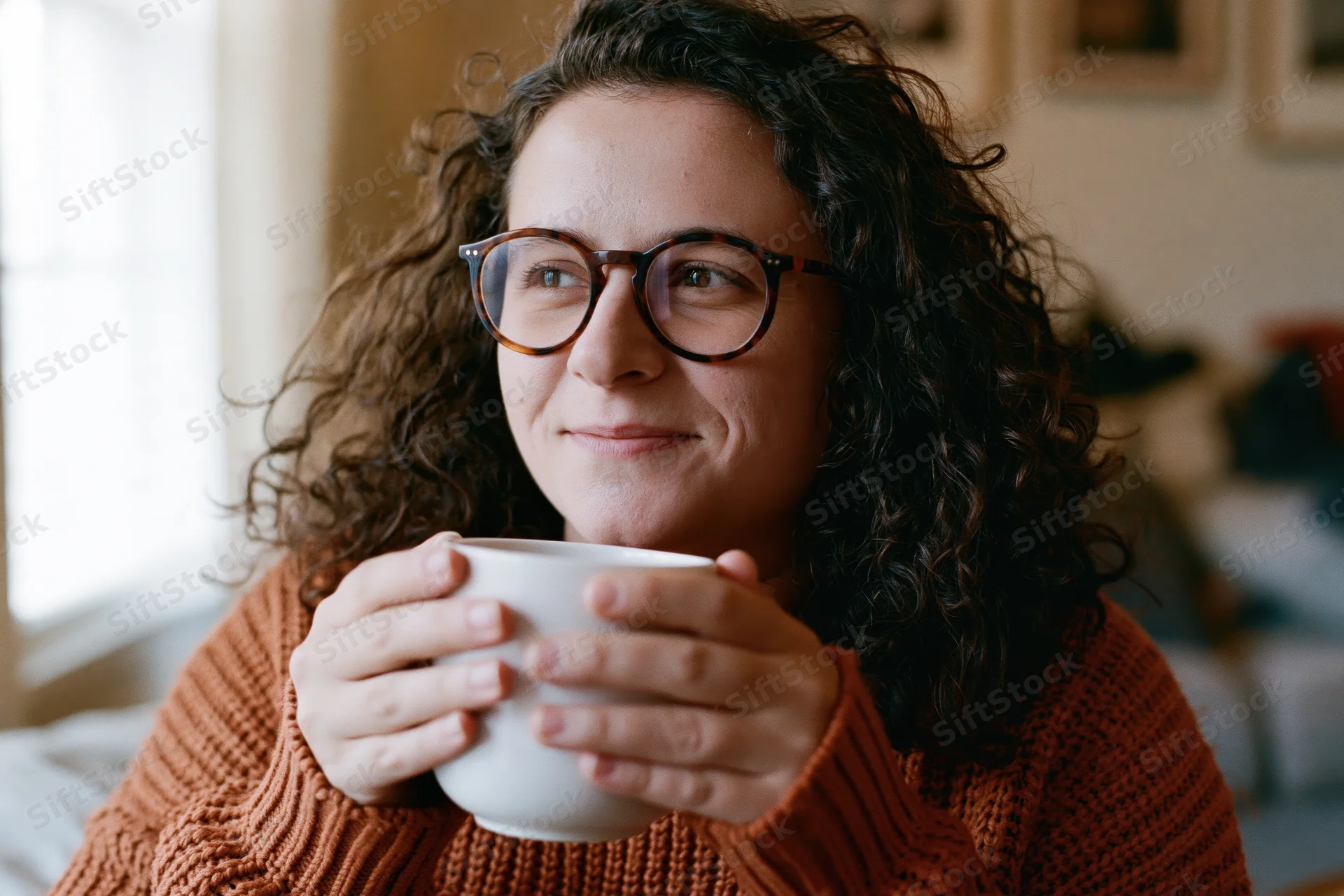 Woman with curly hair and glasses smiling softly while holding a white mug indoors