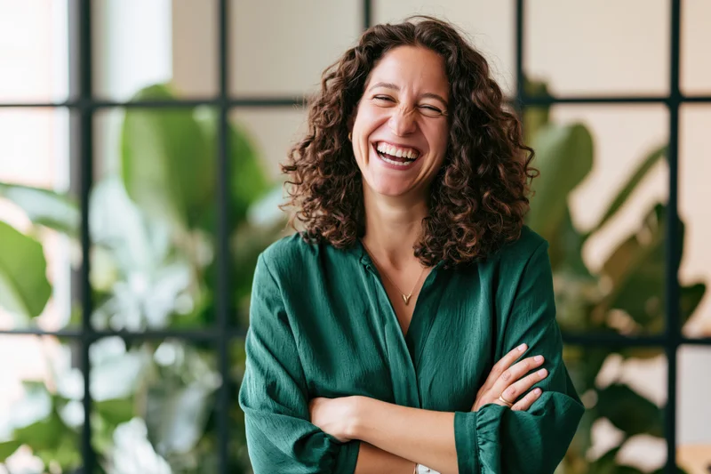 Woman laughing with arms crossed in office interior with plants and window grid