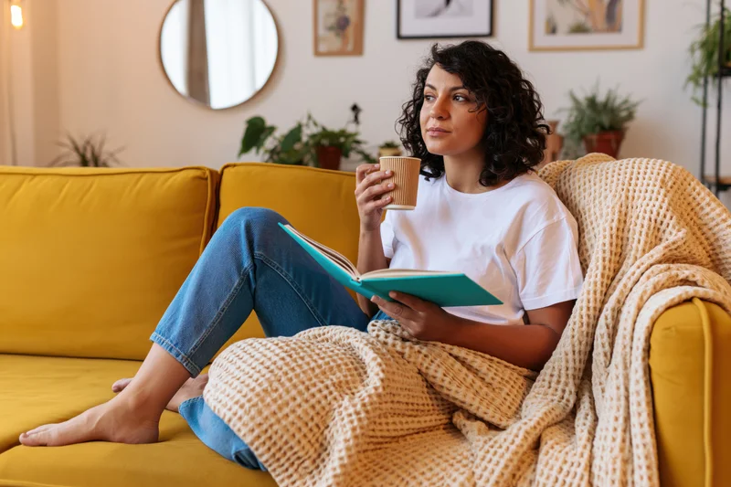 Woman reading a book while holding a coffee mug on a yellow couch at home