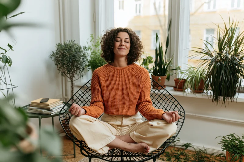 Woman meditating cross-legged in a plant-filled living room by a window