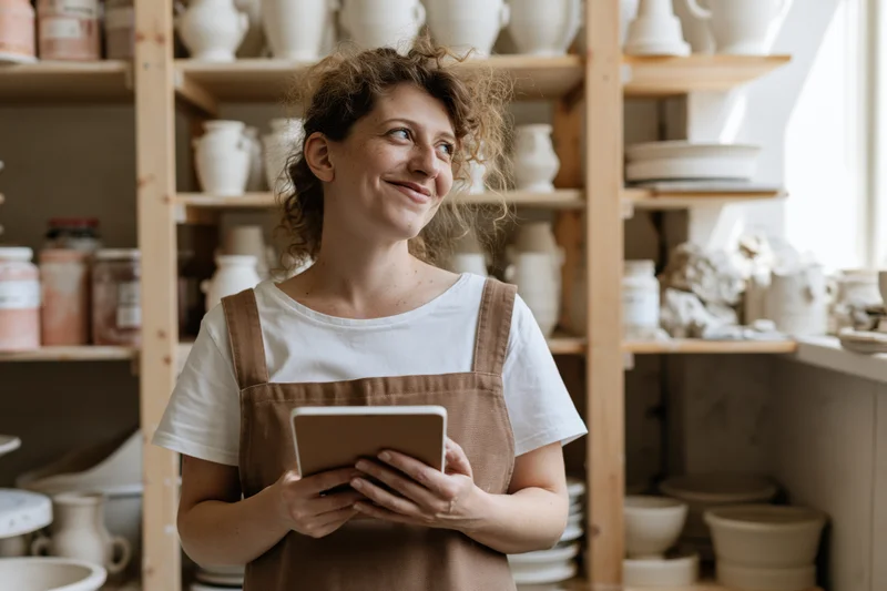 Woman in apron holding tablet in pottery studio with shelves of ceramics behind her