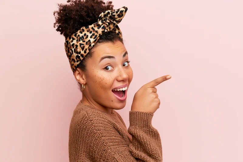 Young woman with curly hair in a bun wearing leopard headband pointing to the side with a happy expression against pink background