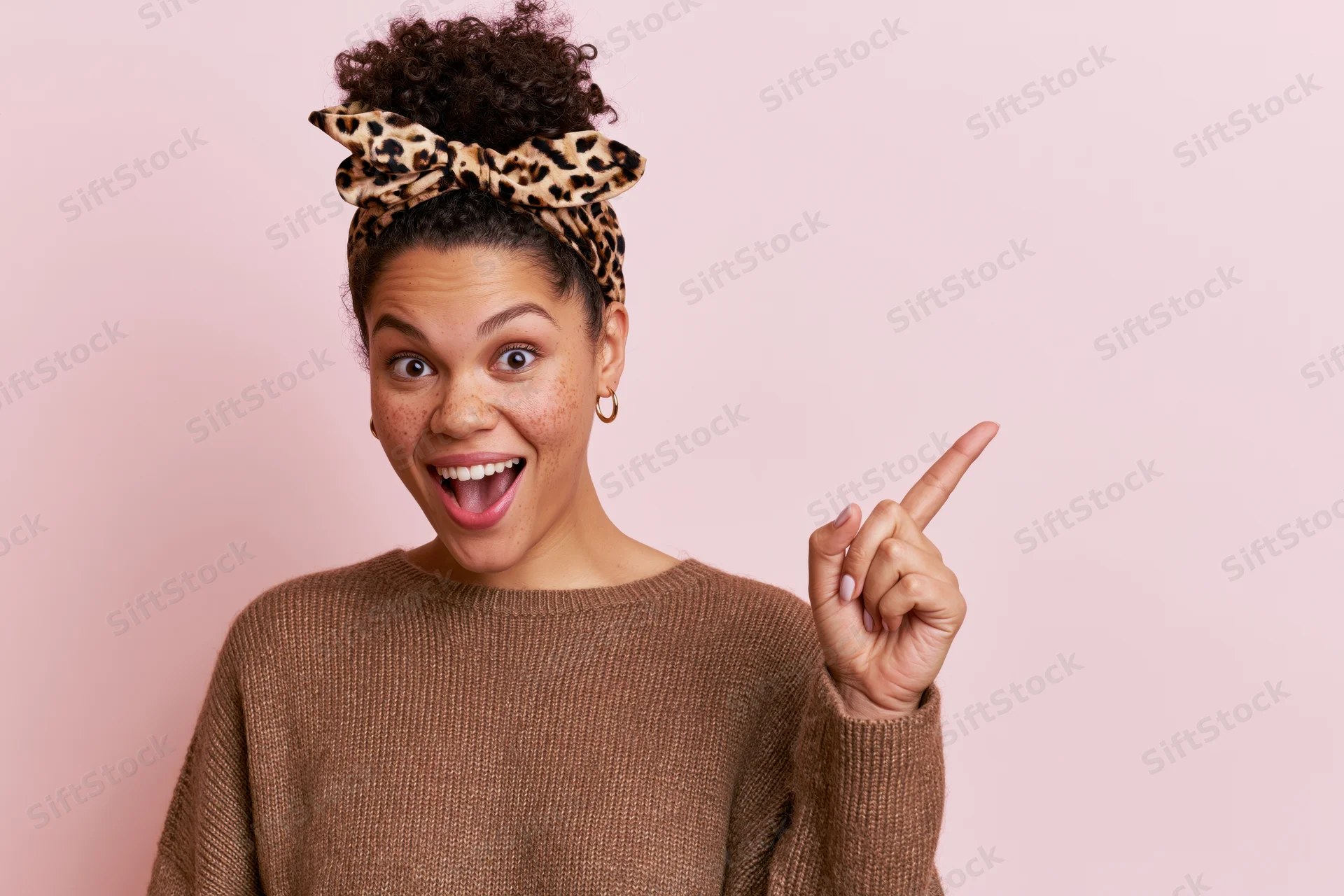 Young woman with curly hair in a bun wearing leopard headband pointing excitedly against pink background