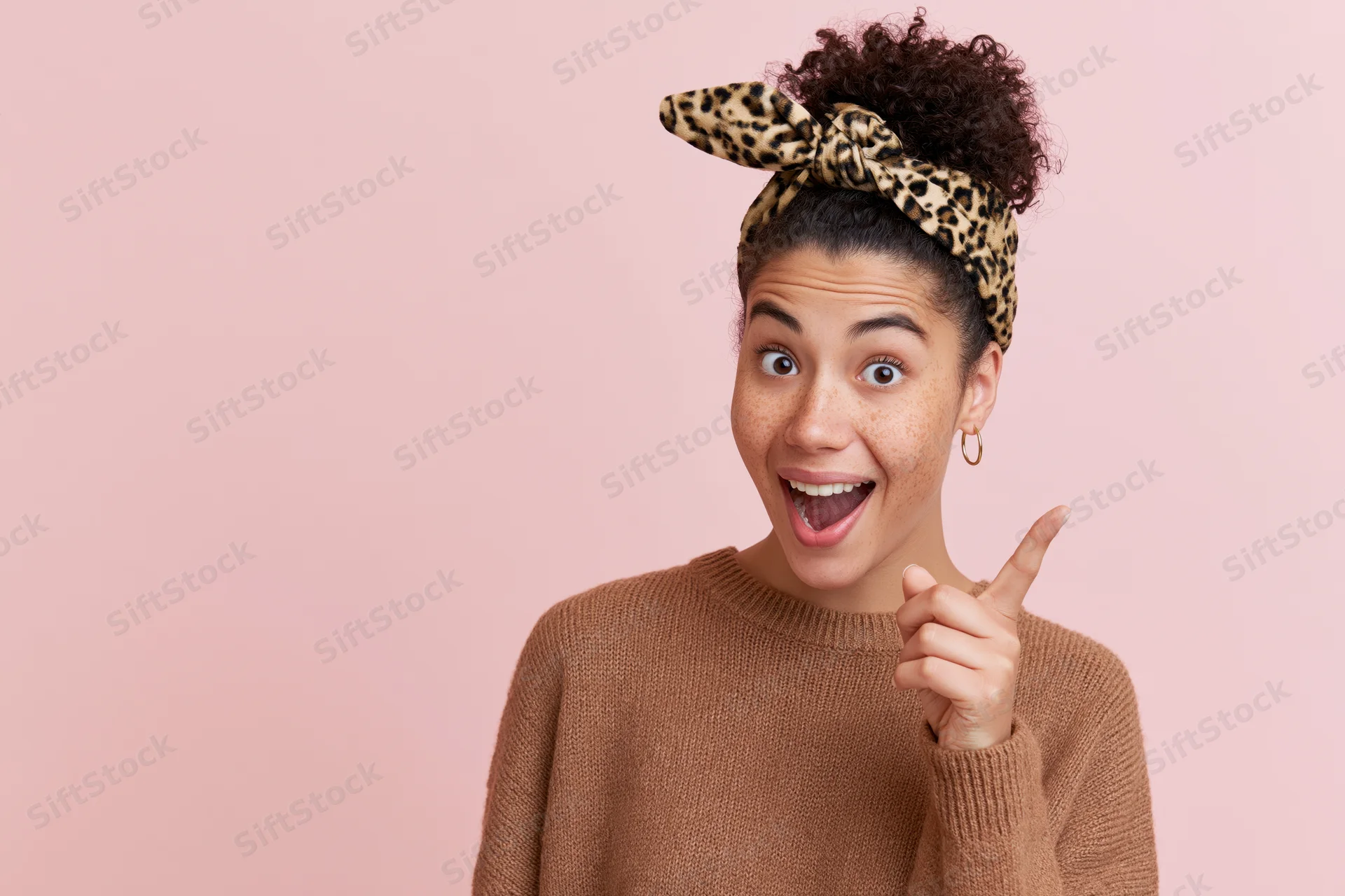 Young woman with curly hair in a high bun wearing a leopard-print headband and brown sweater, pointing excitedly against a pink background