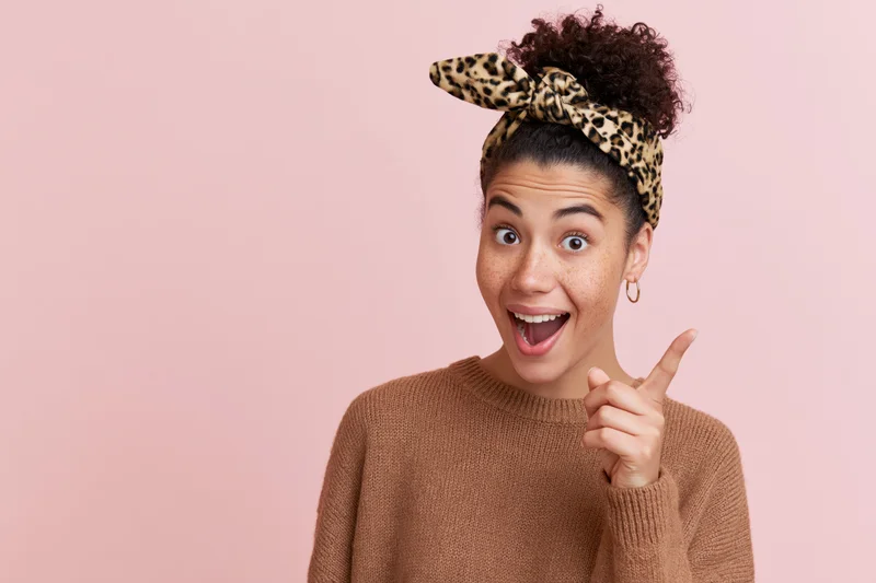 Young woman with curly hair in a high bun wearing a leopard-print headband and brown sweater, pointing excitedly against a pink background