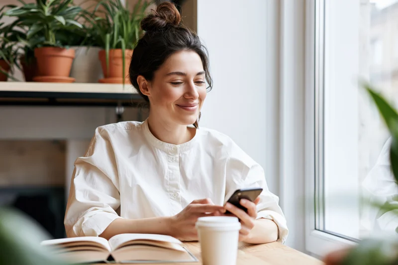 Woman smiling while using smartphone and reading book at wooden table near window