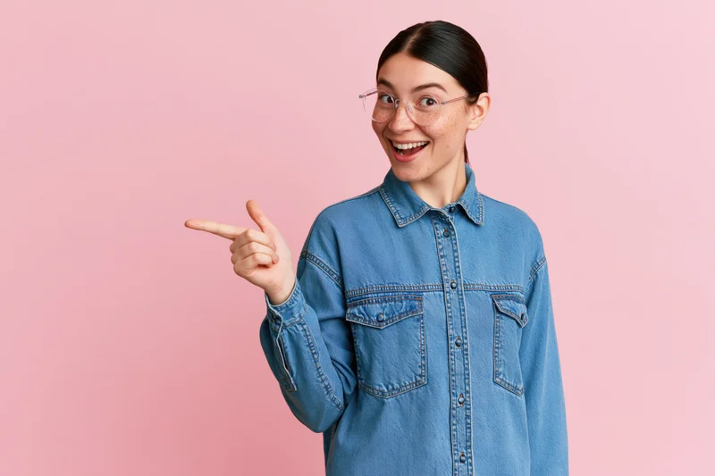 Young woman in denim shirt pointing with smile against pink background