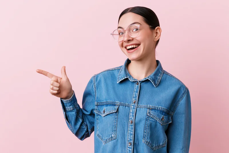 Young woman in denim jacket pointing with smile against pink background