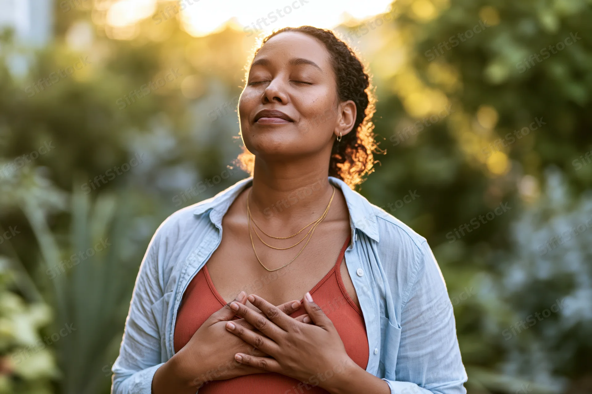 Young black woman with eyes closed and hands on chest in a garden at golden hour