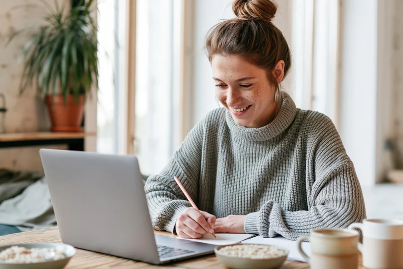 Woman smiling while writing notes with pencil beside laptop at wooden desk in home office
