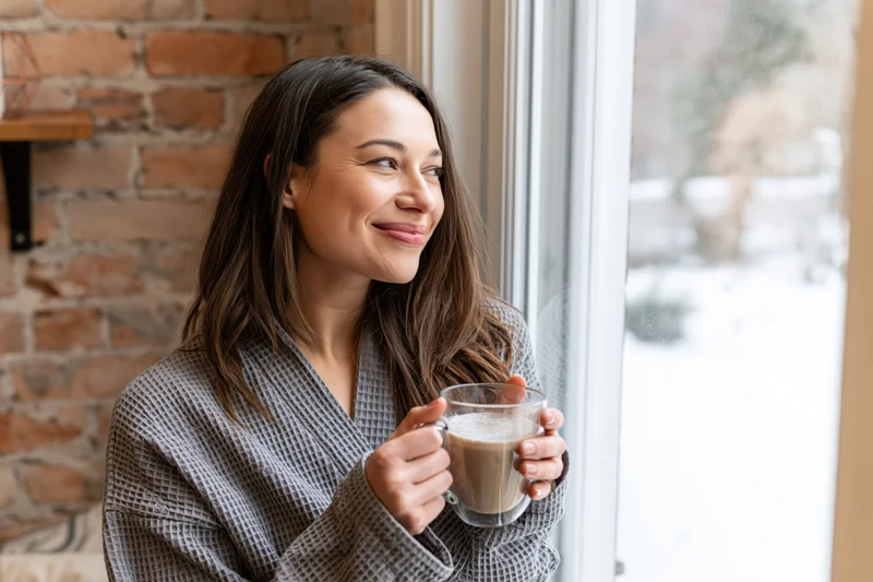 Woman holding glass mug of latte while smiling and looking out snowy window in cozy home setting