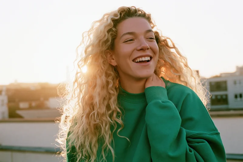 Young woman laughing with long curly blonde hair on rooftop at sunset