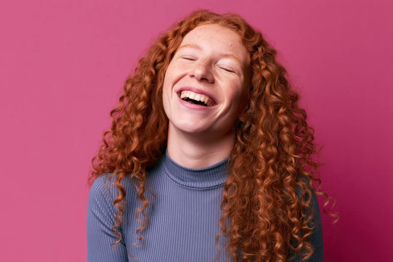 Young woman with long curly red hair laughing wholeheartedly against pink background