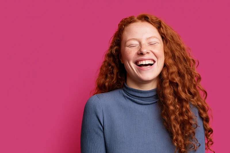 Young woman with long curly red hair laughing wholeheartedly against a pink background