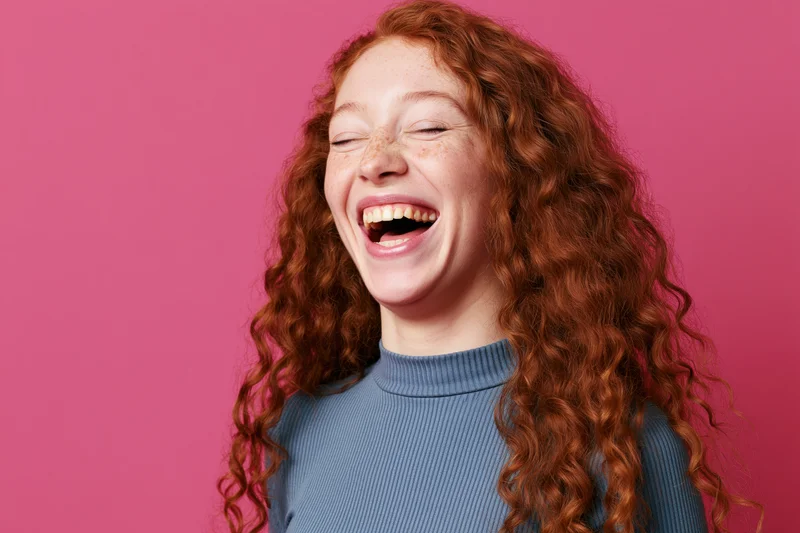 Young woman with long curly red hair laughing wholeheartedly against pink background