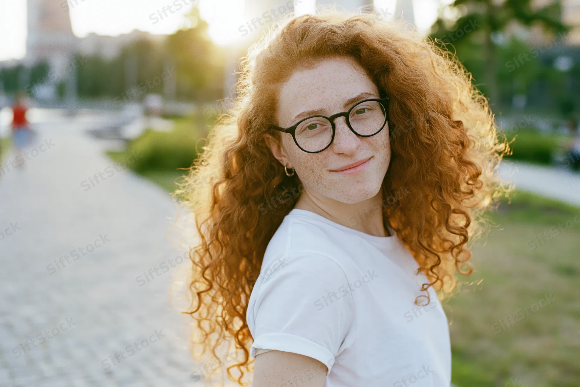Young woman with long red curly hair and glasses smiling over her shoulder in a park at golden hour