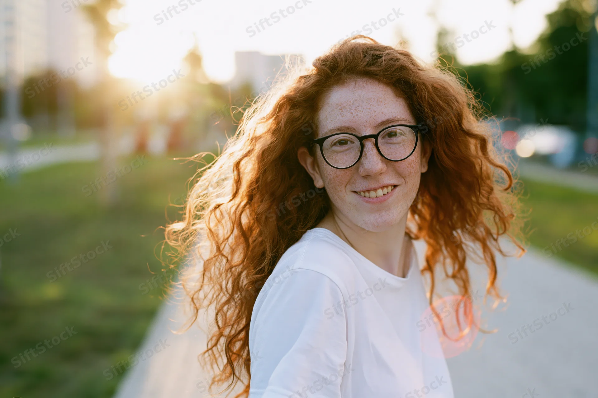 Young woman with long red curly hair smiling while turning over her shoulder on a park pathway at golden hour
