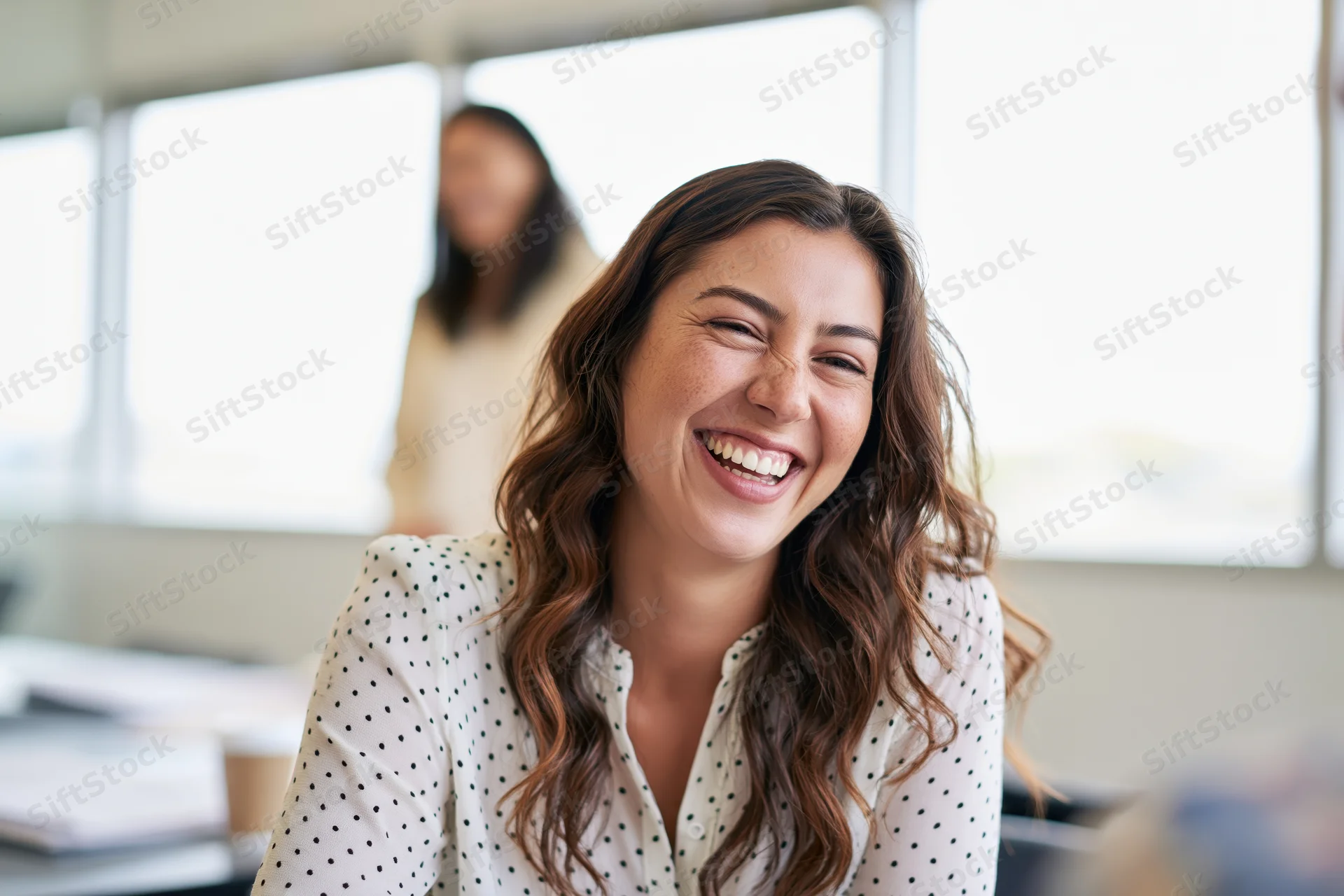 Young woman laughing in an office setting with a colleague in the background