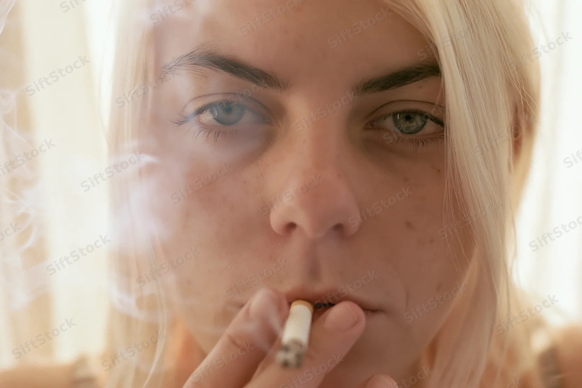 Young woman smoking a cigarette indoors, close-up on her face with smoke swirling around her eyes