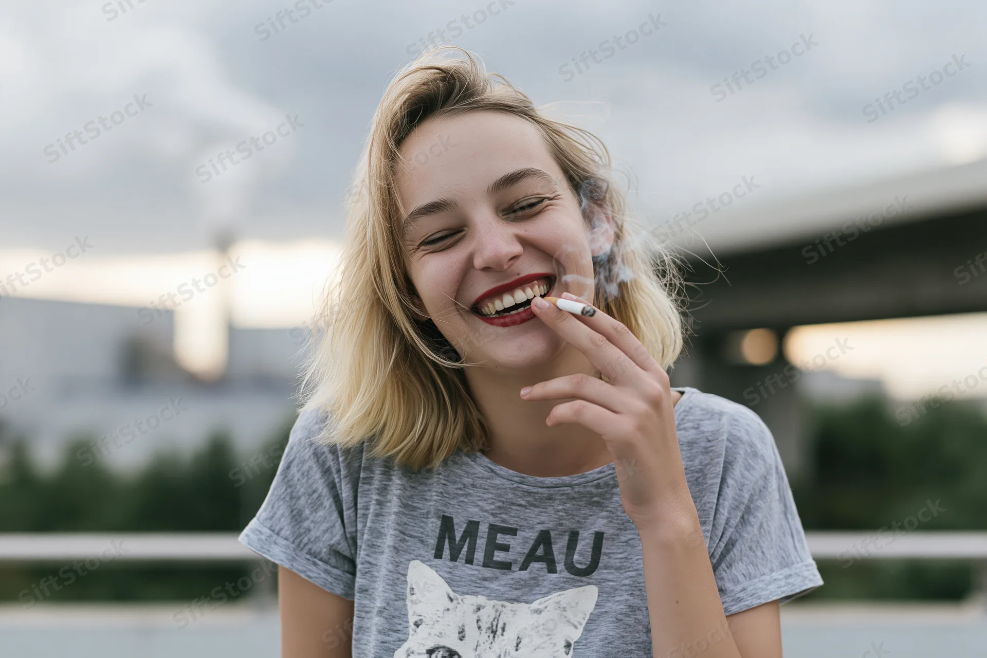 Young woman laughing while smoking a cigarette outdoors near a bridge