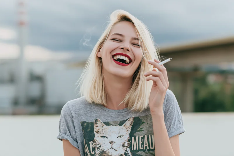 Young woman laughing with cigarette in hand outdoors near bridge