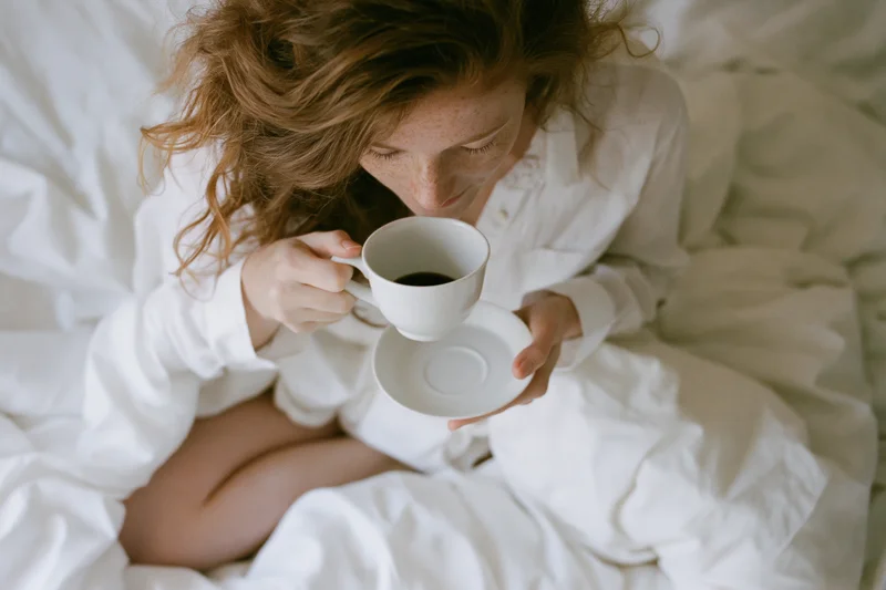 Woman drinking coffee from a white cup and saucer while sitting cross-legged in bed