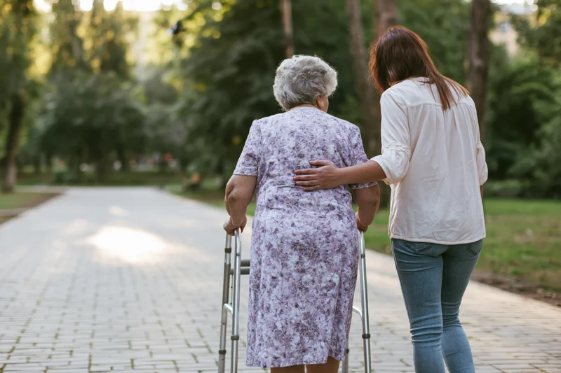 Elderly woman using walker supported by younger woman walking in park