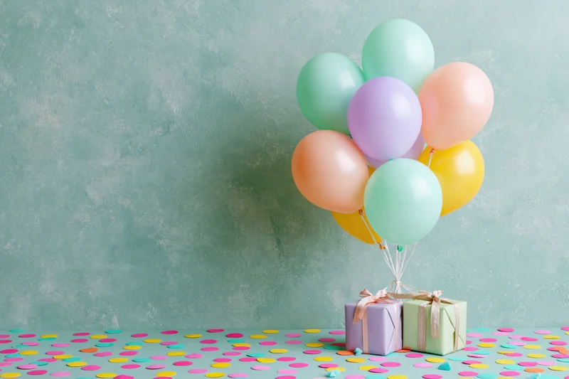 Pastel balloons tied to gift boxes on a confetti-covered surface against a textured wall