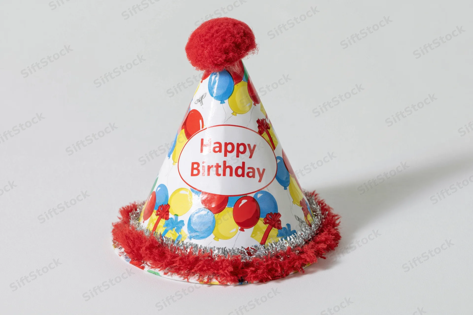 Colorful paper birthday party hat with red pom-pom and balloon pattern sitting upright on white surface