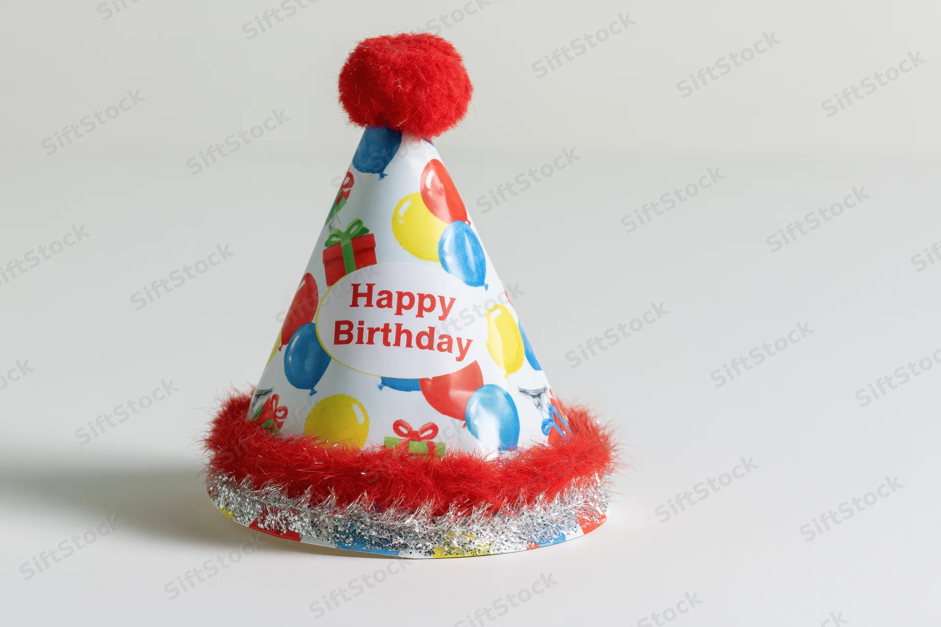 Colorful paper birthday party hat with red pompon and tinsel trim standing upright on white surface