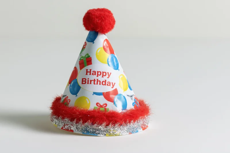 Colorful paper birthday party hat with red pompon and tinsel trim standing upright on white surface