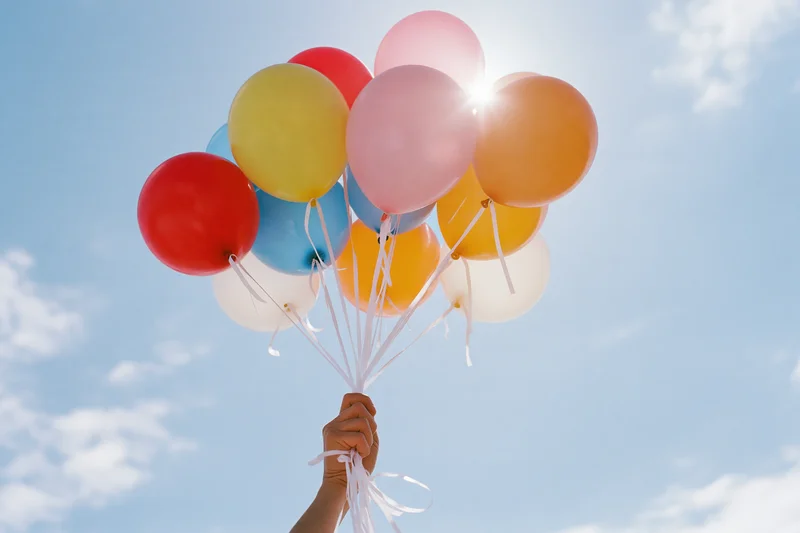 Hand holding a bunch of multicolored latex balloons against a blue sky with sunlight and clouds