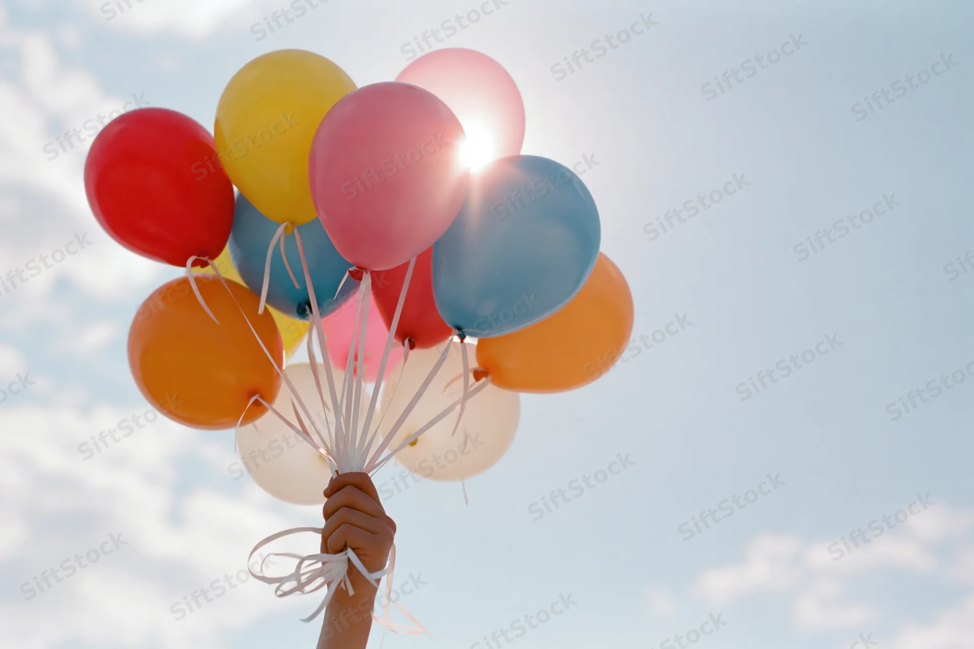 Hand holding a bunch of colorful latex balloons against a bright sky with sun shining through