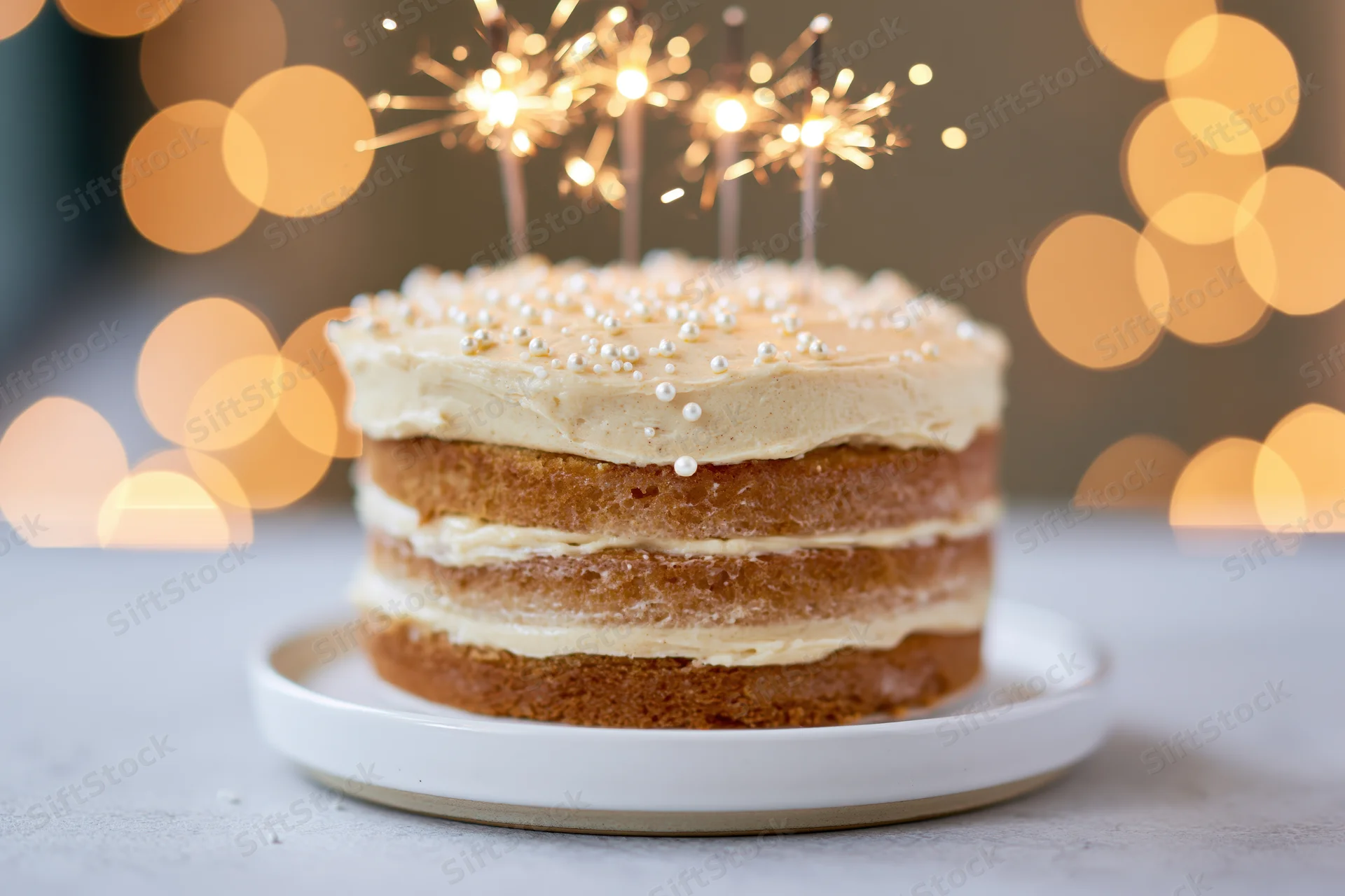 Layered birthday cake with sparklers on a white ceramic stand against warm bokeh lights