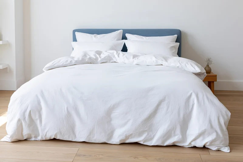 Neatly made bed with white cotton duvet and multiple white pillows against a blue headboard in a minimalist bedroom