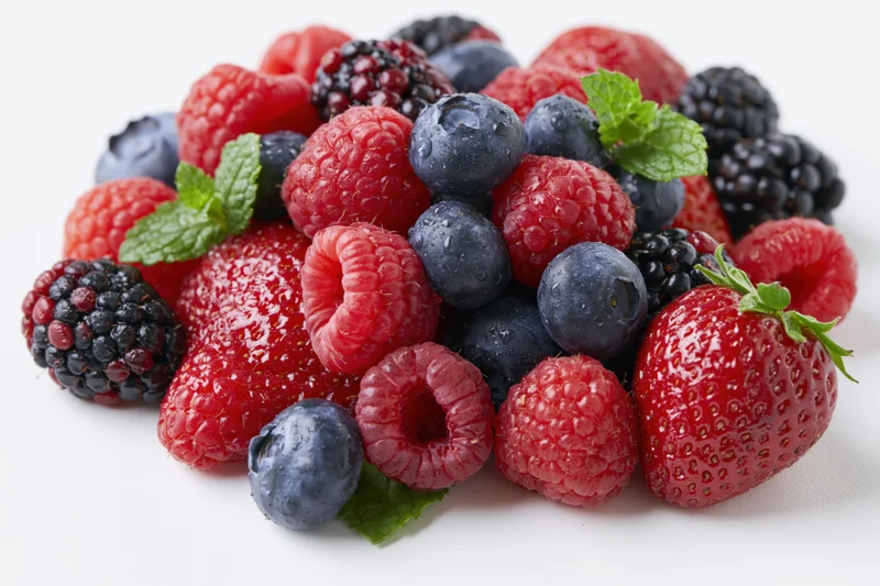 A pile of mixed fresh berries including raspberries, blueberries, strawberries, and blackberries with mint leaves on a white surface