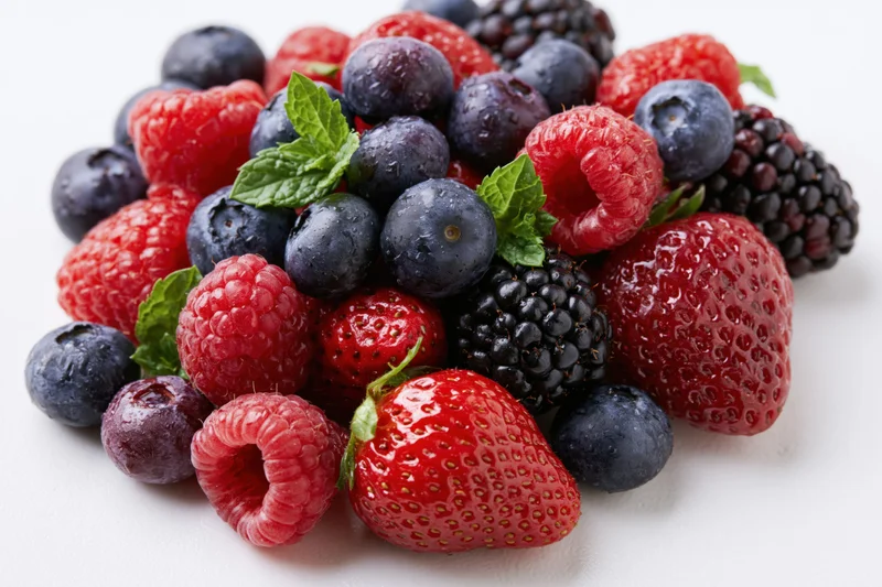 A pile of mixed fresh berries including strawberries, raspberries, blueberries and blackberries on a white surface