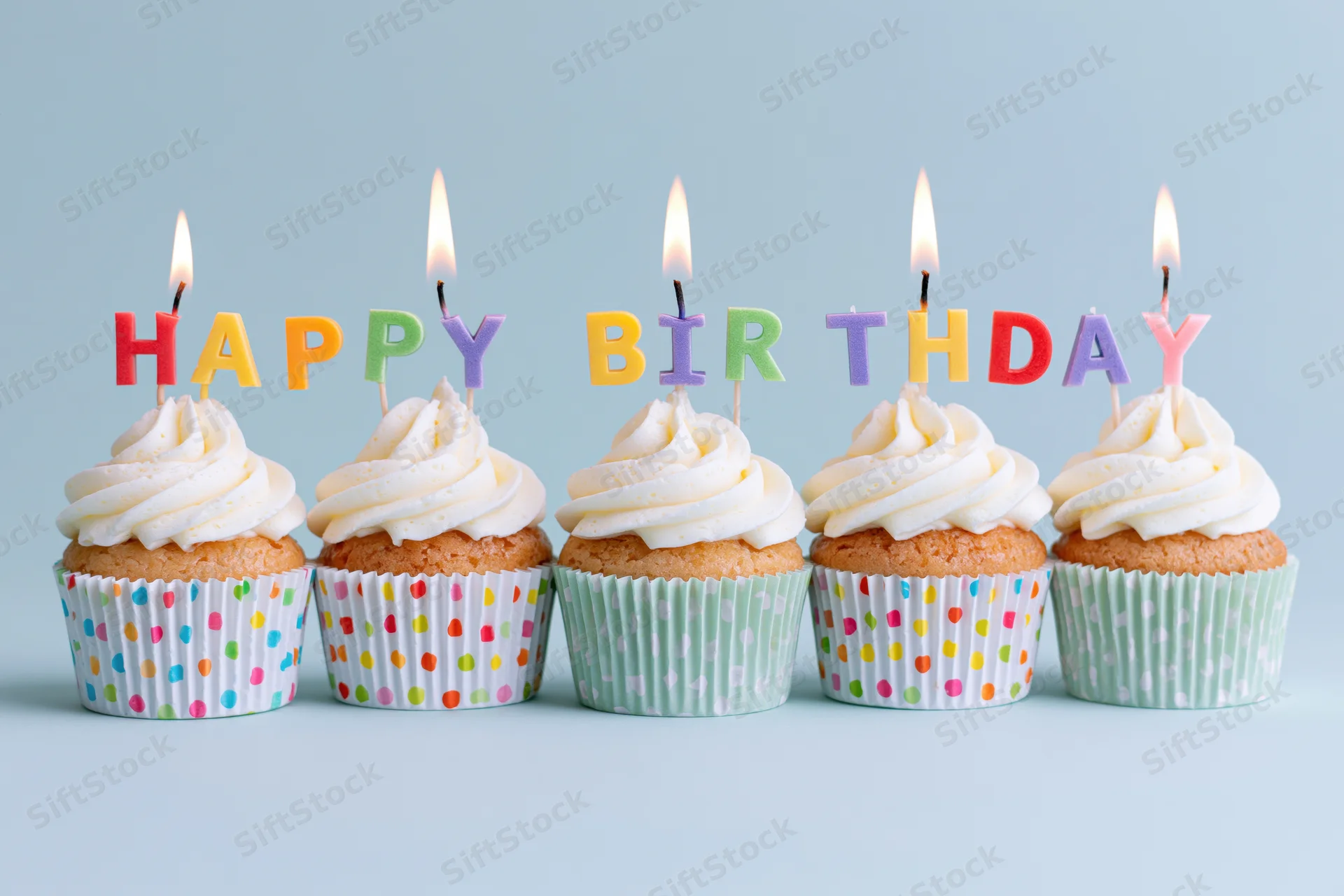 Five cupcakes with white frosting and lit letter candles spelling happy birthday in colorful wrappers on a light blue surface